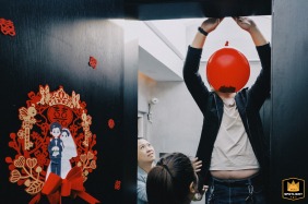 In Kaifeng, China, friends help hang balloons, but one accidentally blocks the subject's head, creating a humorous, coincidental moment during the wedding preparations.
